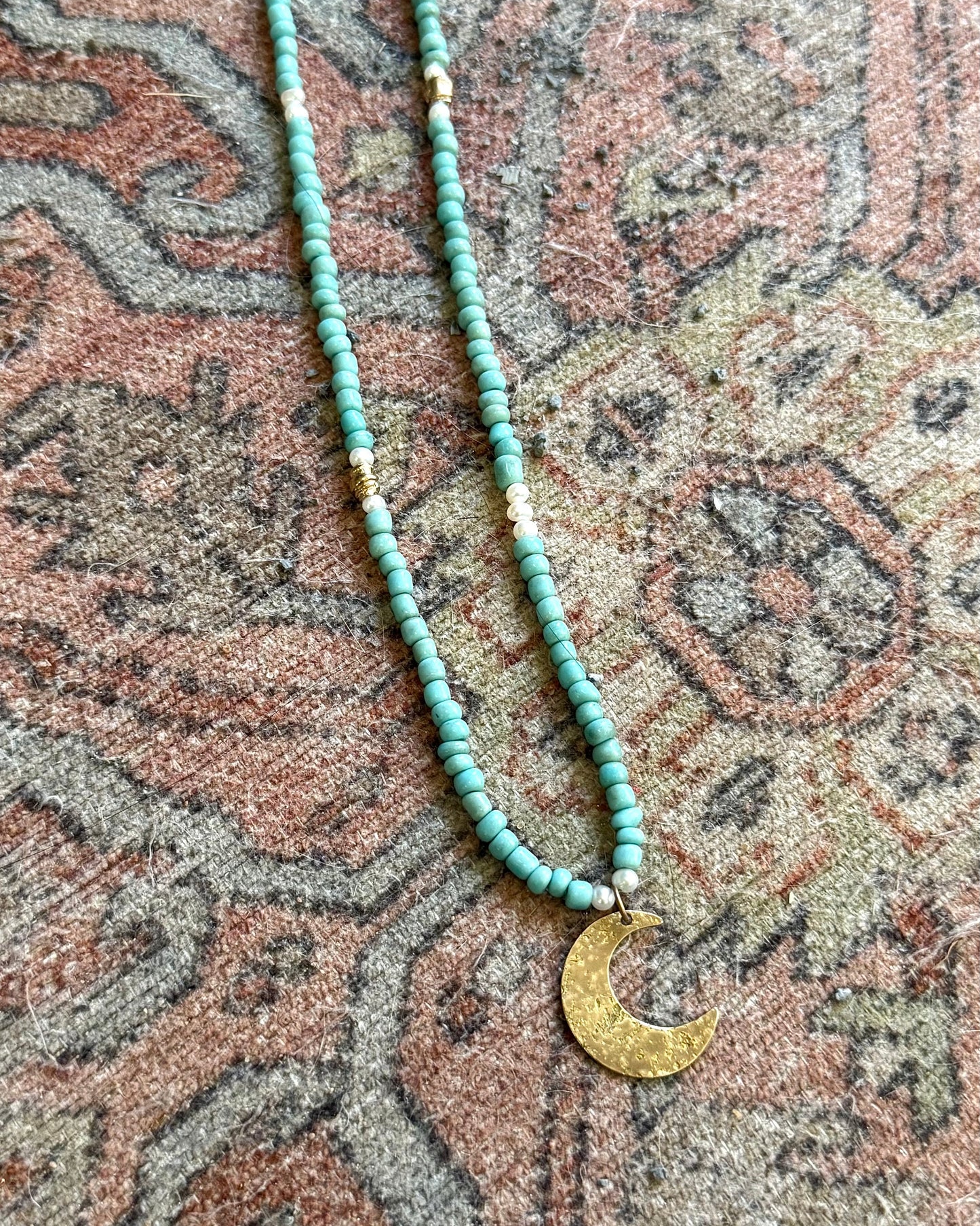 Turquoise beaded necklace with a gold crescent moon pendant on a patterned fabric background