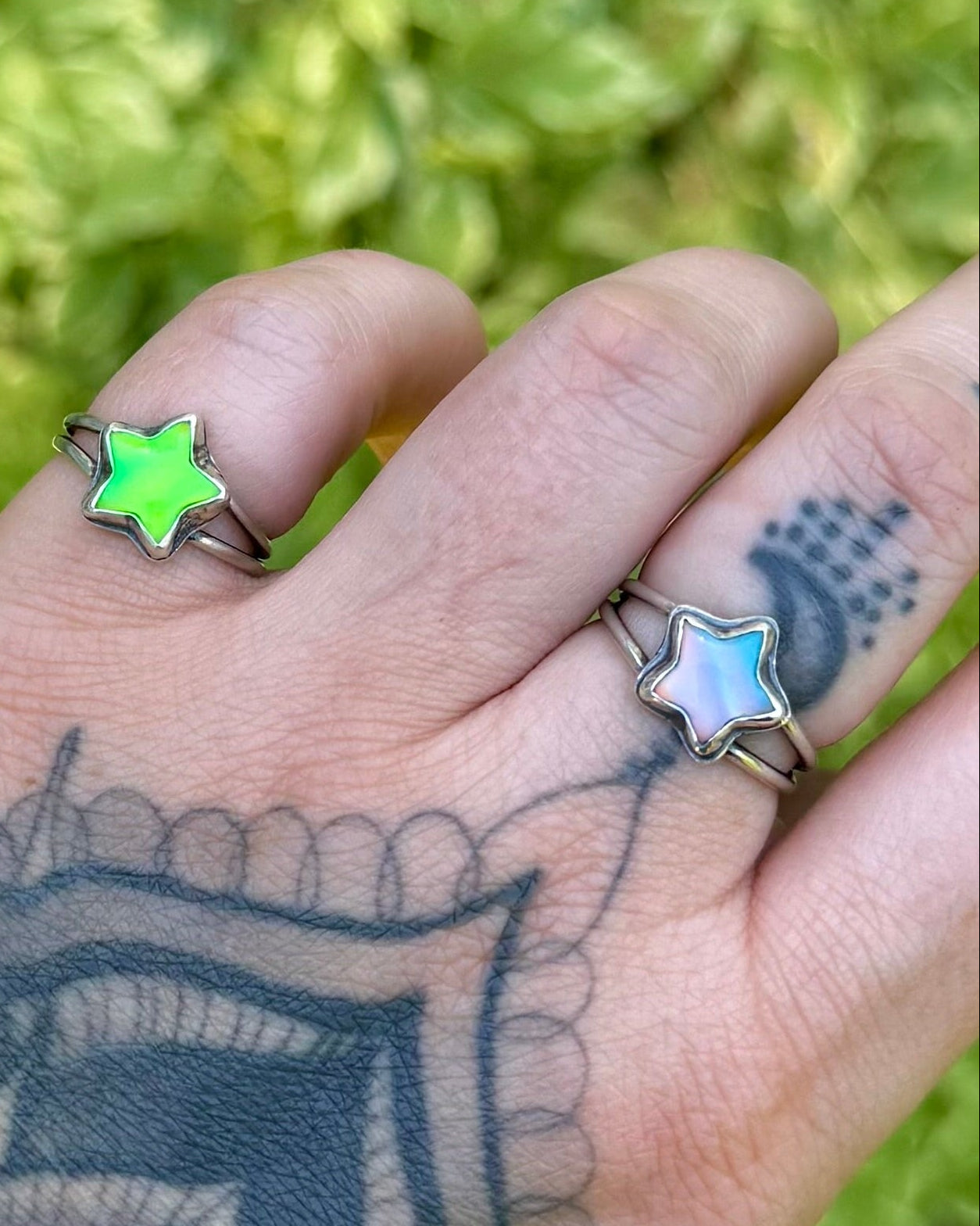 Hand with tattoos and rings against a green leafy background