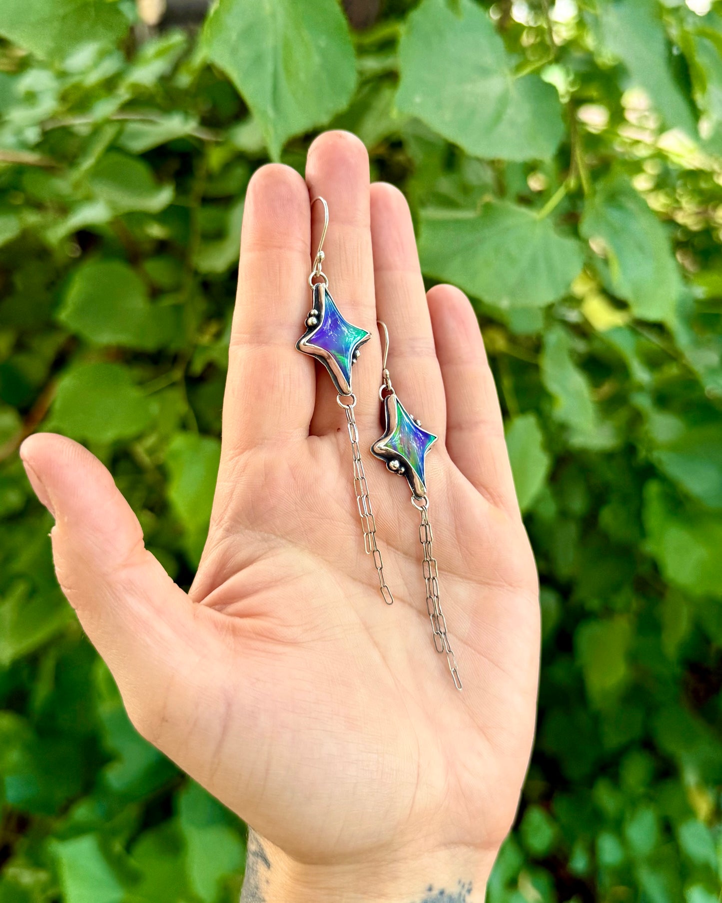 A person's hand holding a pair of dangling star-shaped earrings with iridescent blue and green enamel and silver-toned chains, set against a background of green leaves.