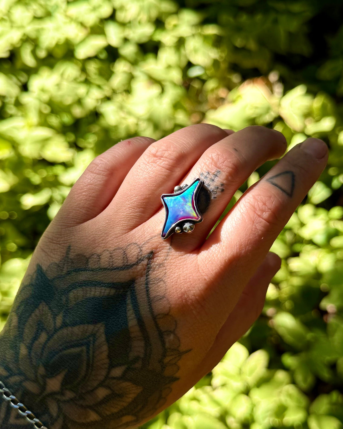 A tattooed hand wearing a silver ring with a blue star-shaped inlay, set against a background of green foliage.