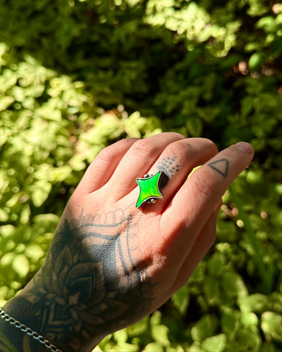 A tattooed hand wearing a bright green star-shaped ring, set against a blurred background of green foliage.