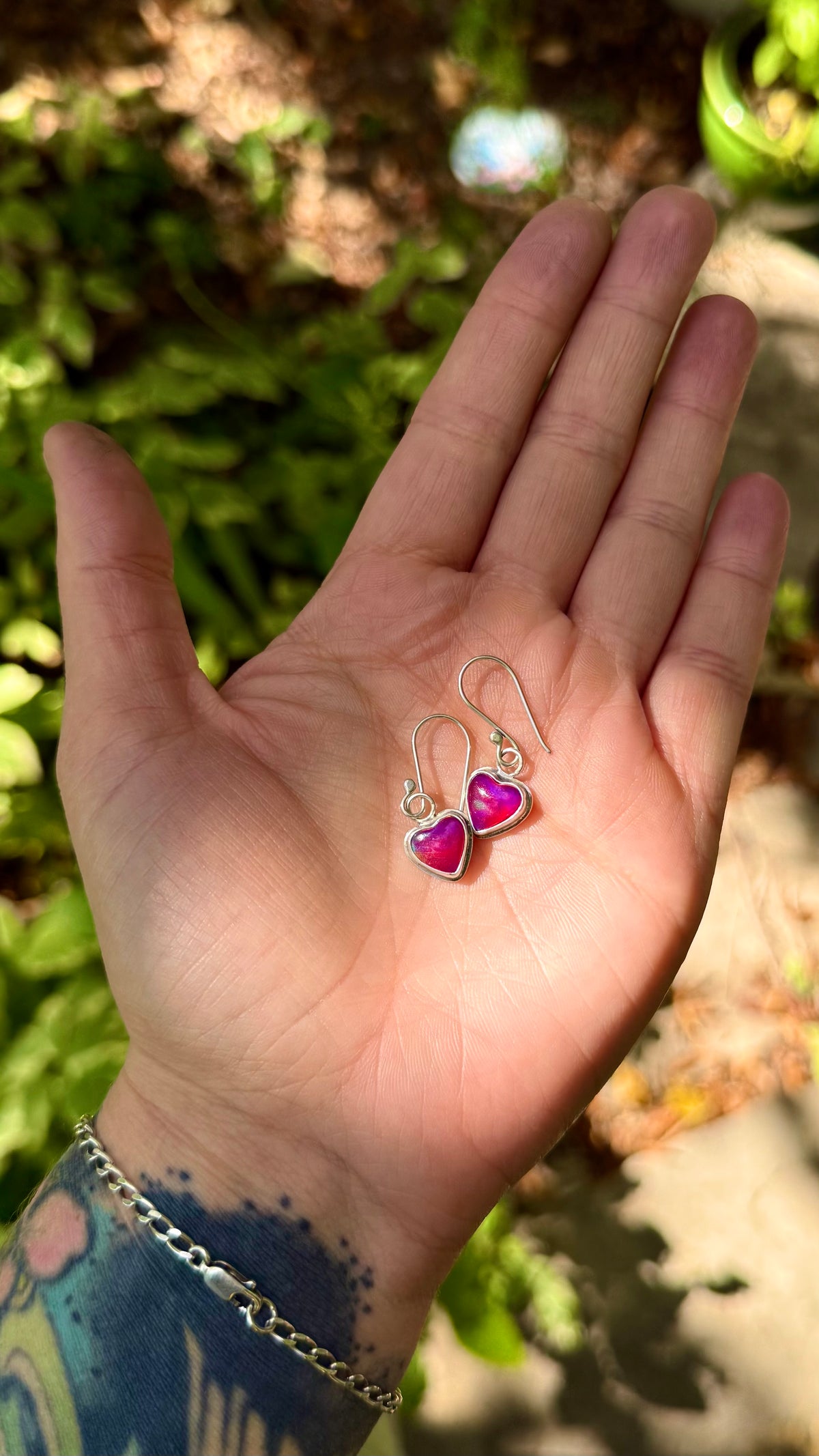 A person's hand holding a pair of silver dangle earrings with pink heart charms, set against a blurred outdoor background of green foliage.