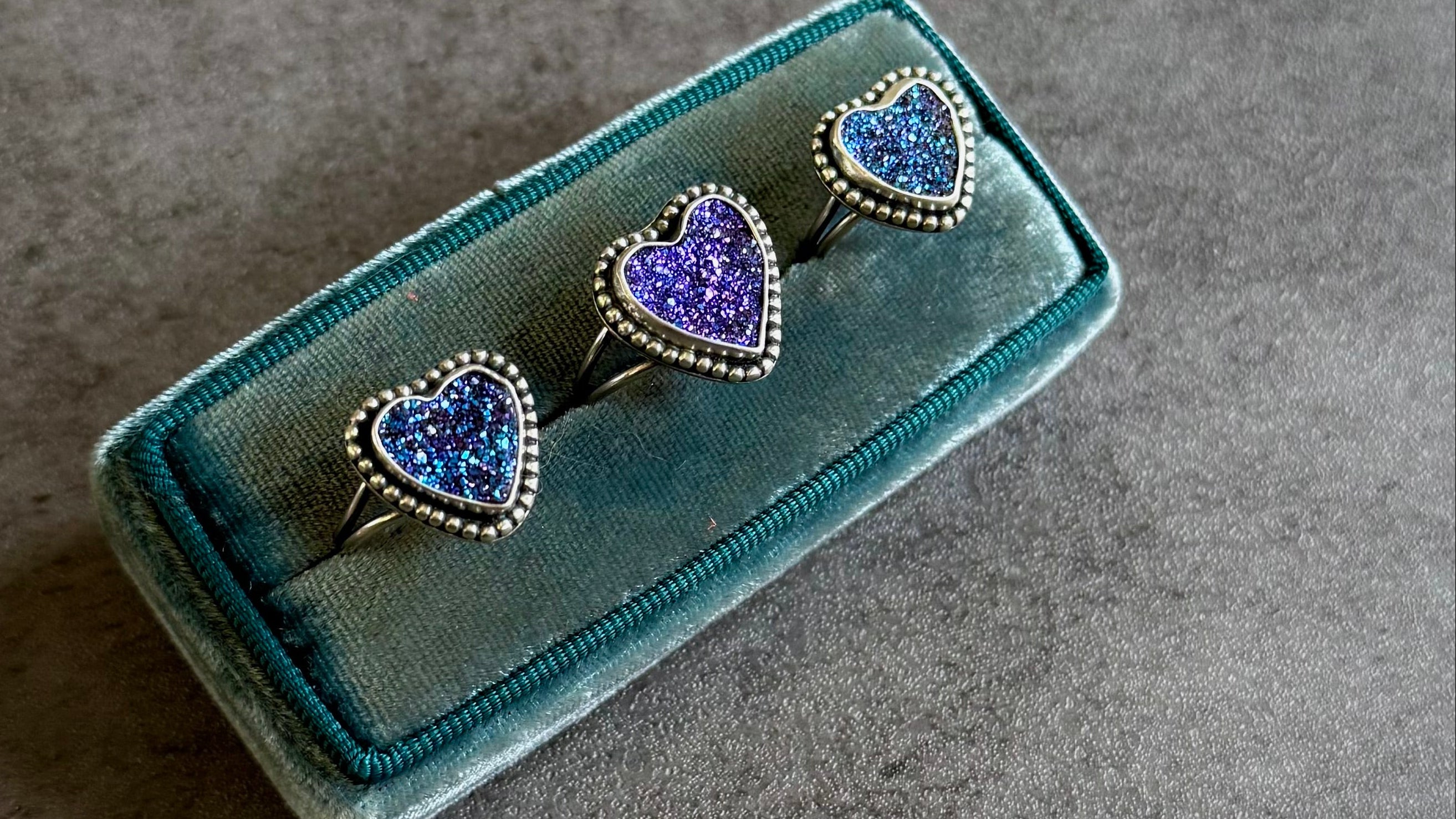 Heart-shaped rings with purple and blue gemstones on a green velvet box against a gray background