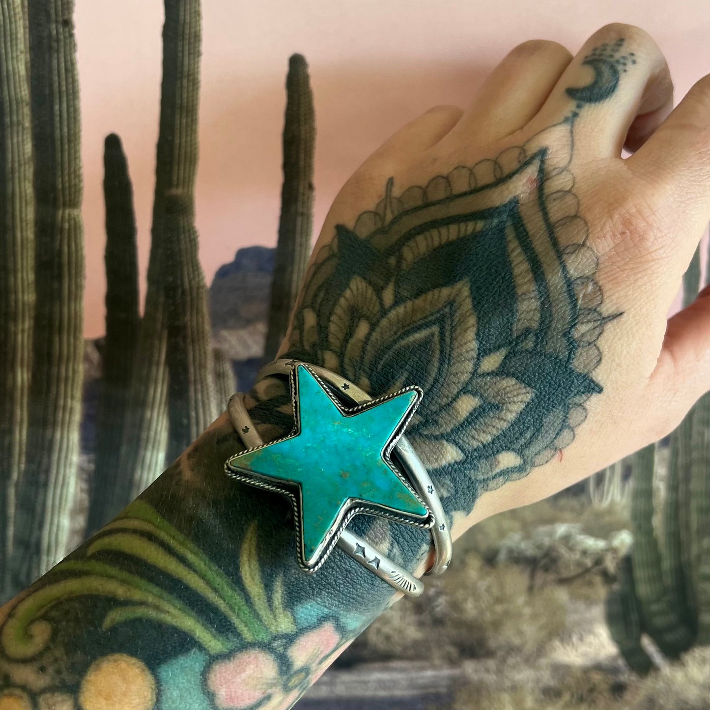 Kingman Turquoise Statement Cuff with a bold turquoise star centerpiece and sterling silver band, styled on a tattooed wrist against a desert cactus background. S/M wrist fit.