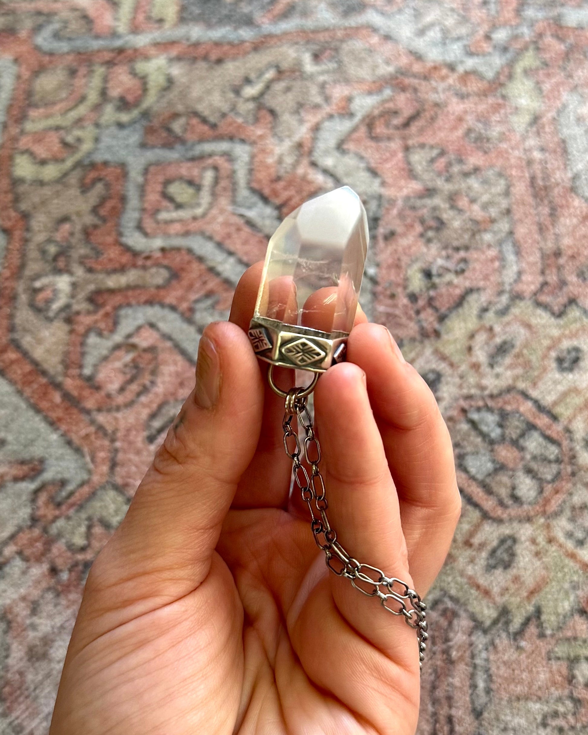 Hand holding a crystal pendant with a chain against a patterned rug background