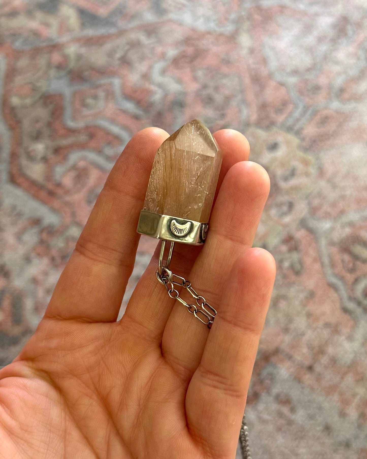 Hand holding a small wooden object with a chain on a patterned rug.