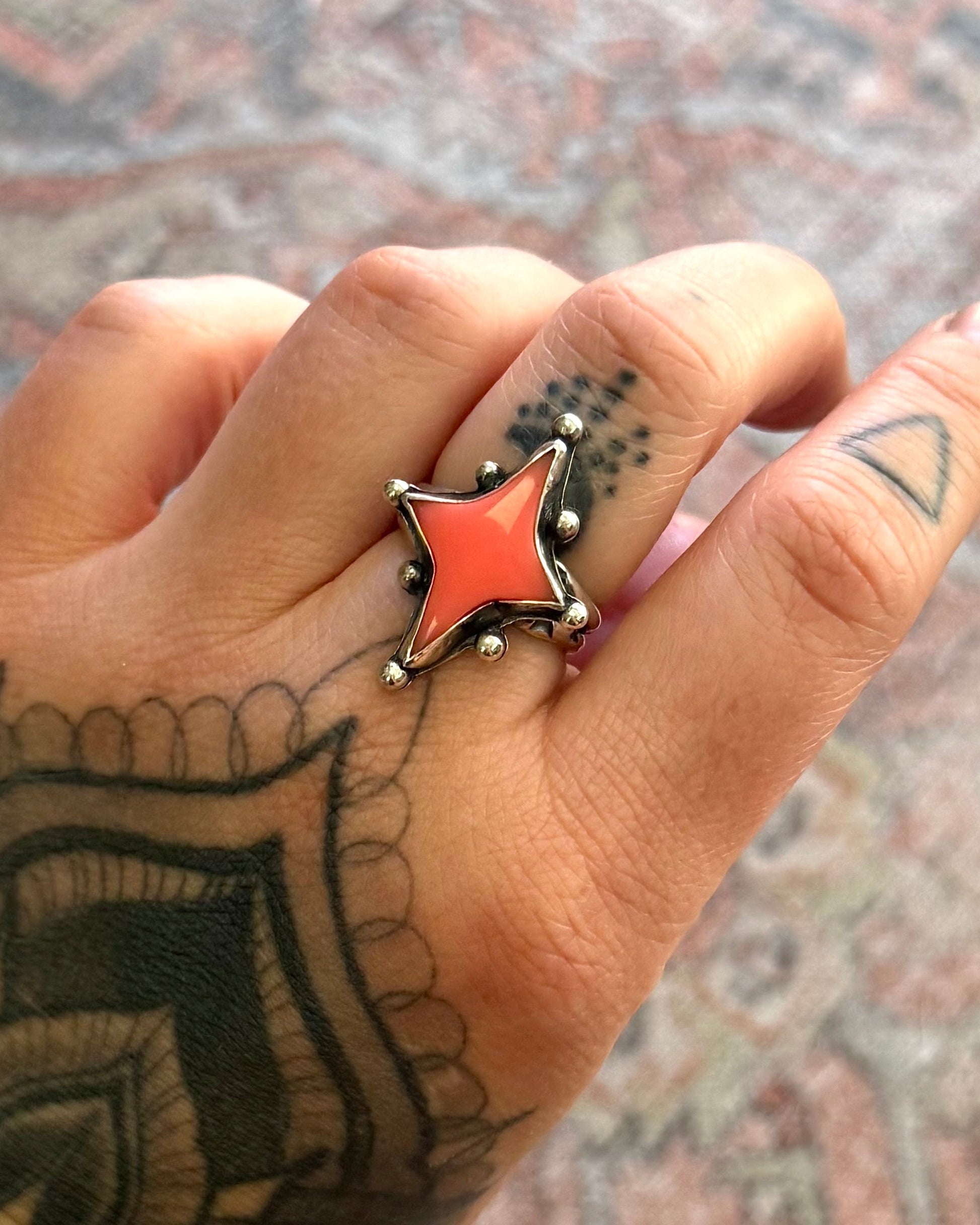 Hand with tattoos wearing a red star-shaped ring on a patterned surface