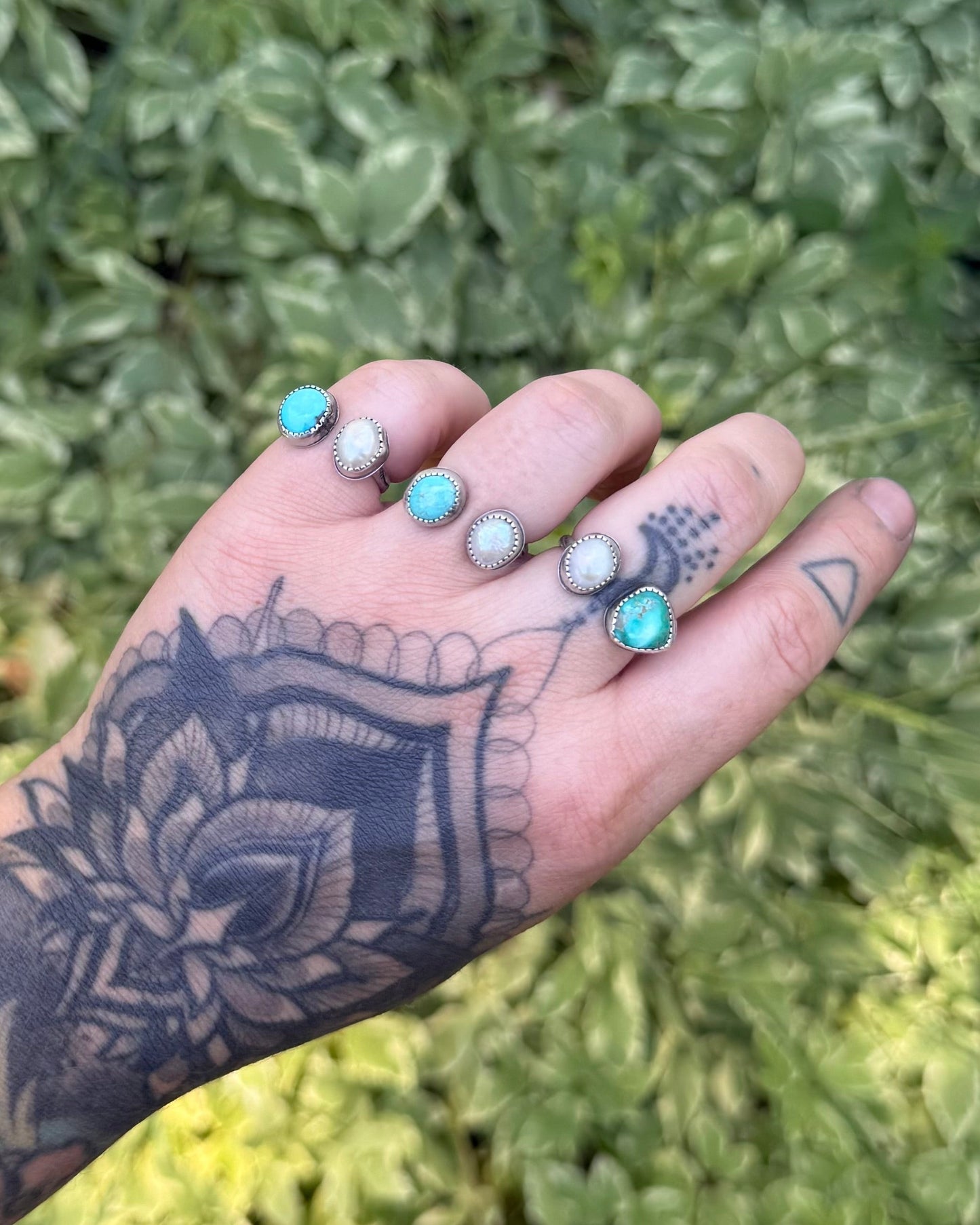 Hand with tattoos and turquoise beads against a green foliage background