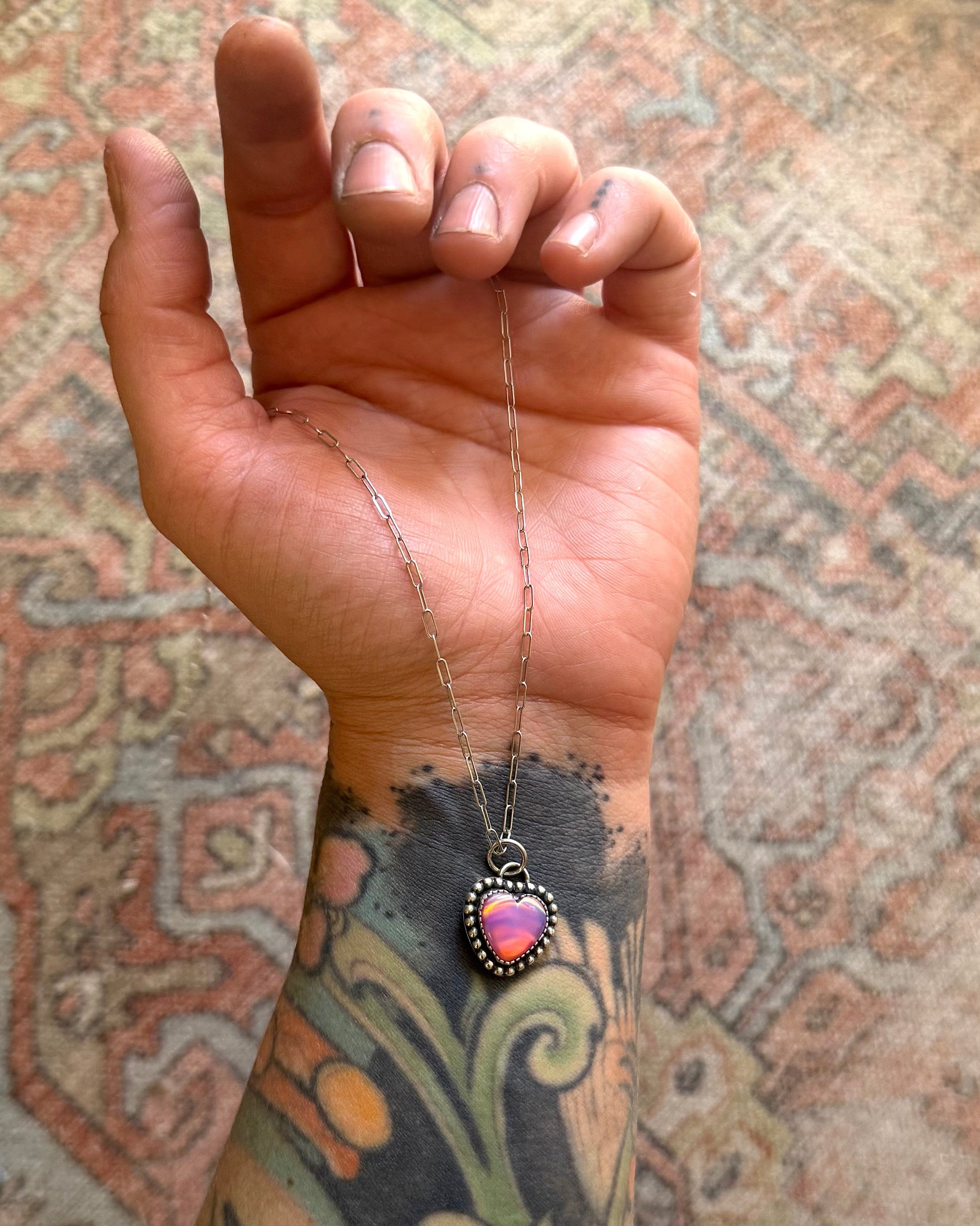 Hand with a tattoo holding a necklace with a heart pendant against a patterned background