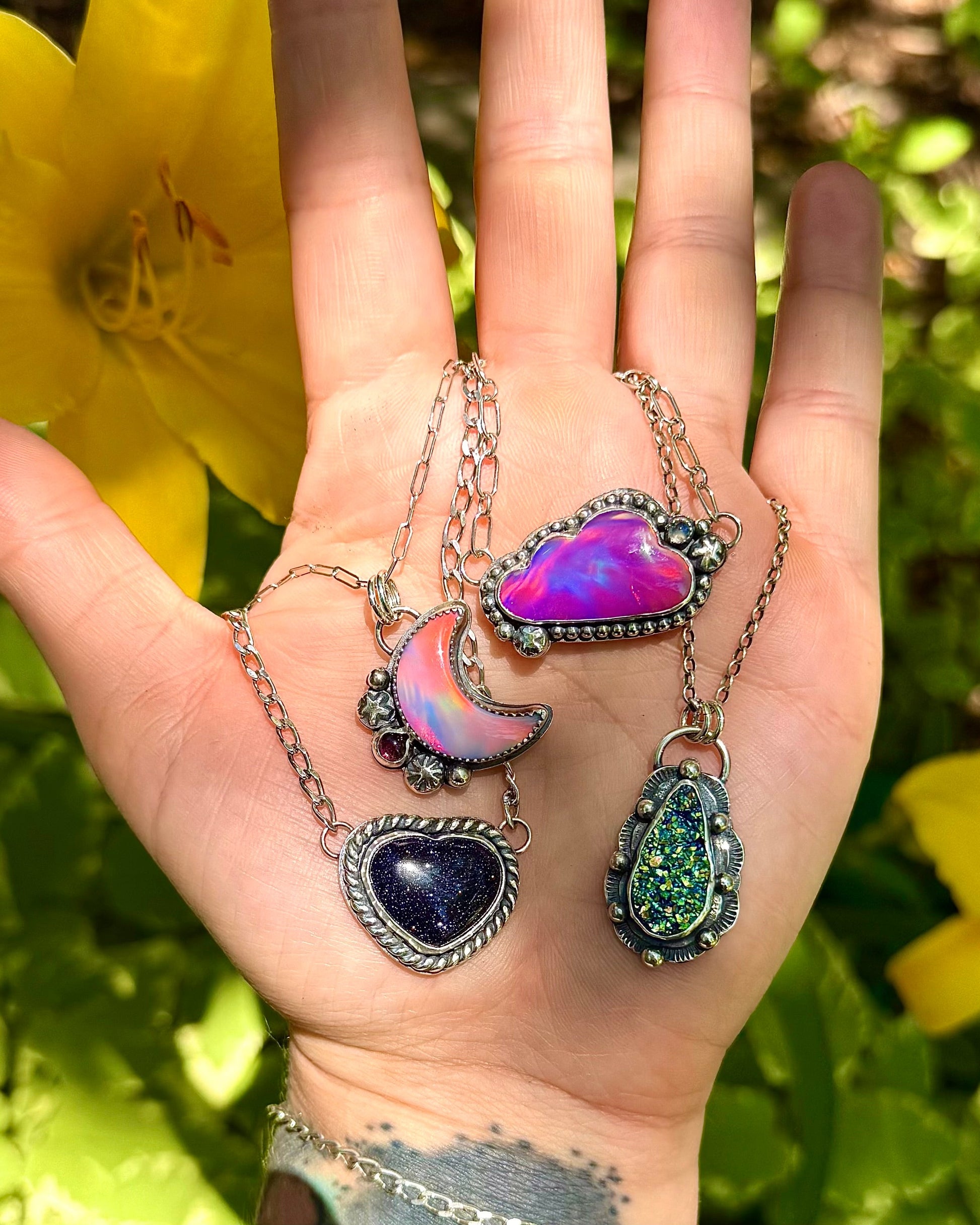 Hand holding four colorful pendant necklaces against a natural background with yellow flowers.