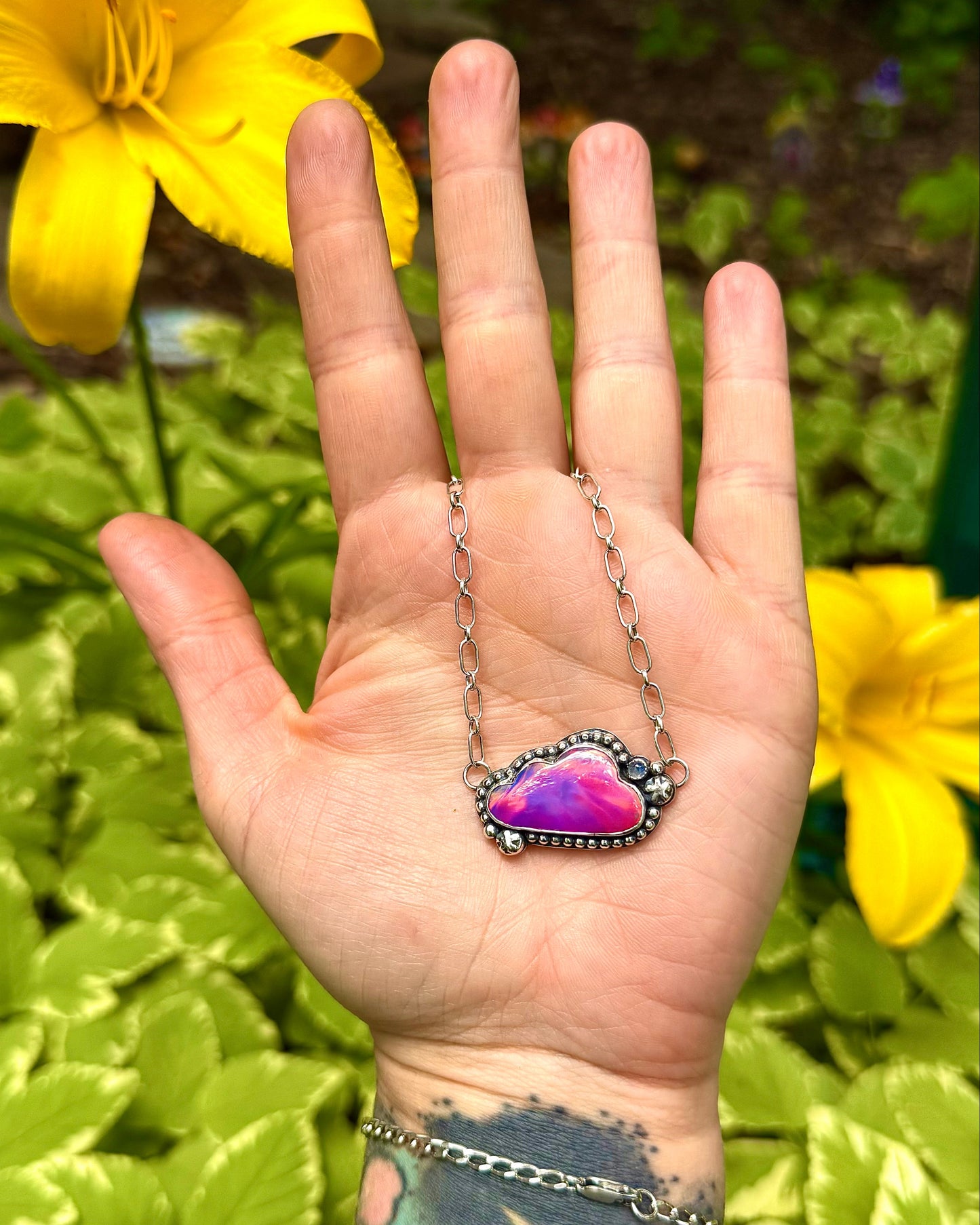 Hand wearing a necklace with an opal pendant against a natural background with yellow flowers.