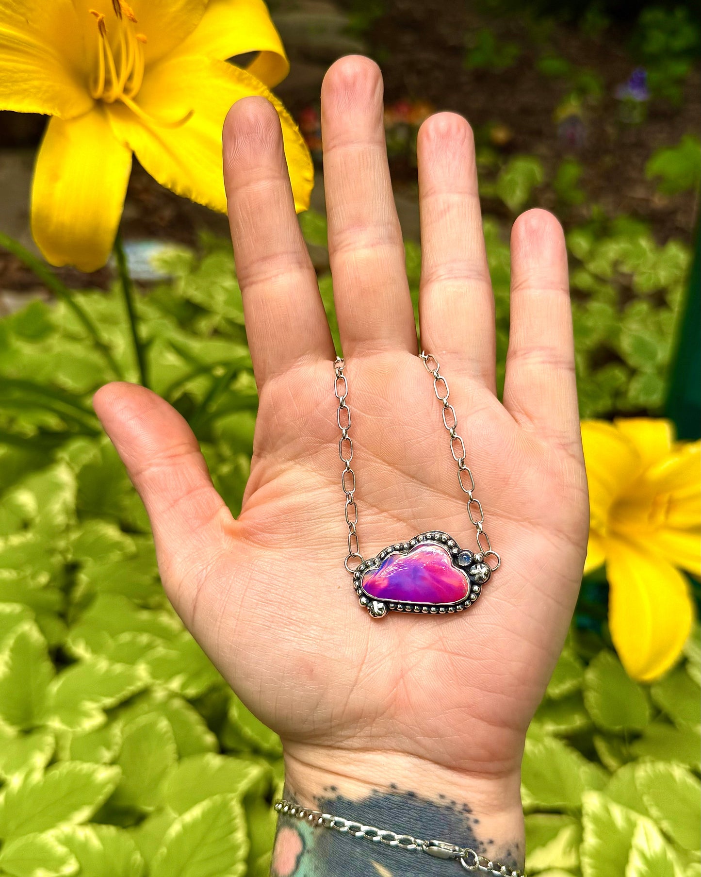 Hand wearing a necklace with an opal pendant against a natural background with yellow flowers.