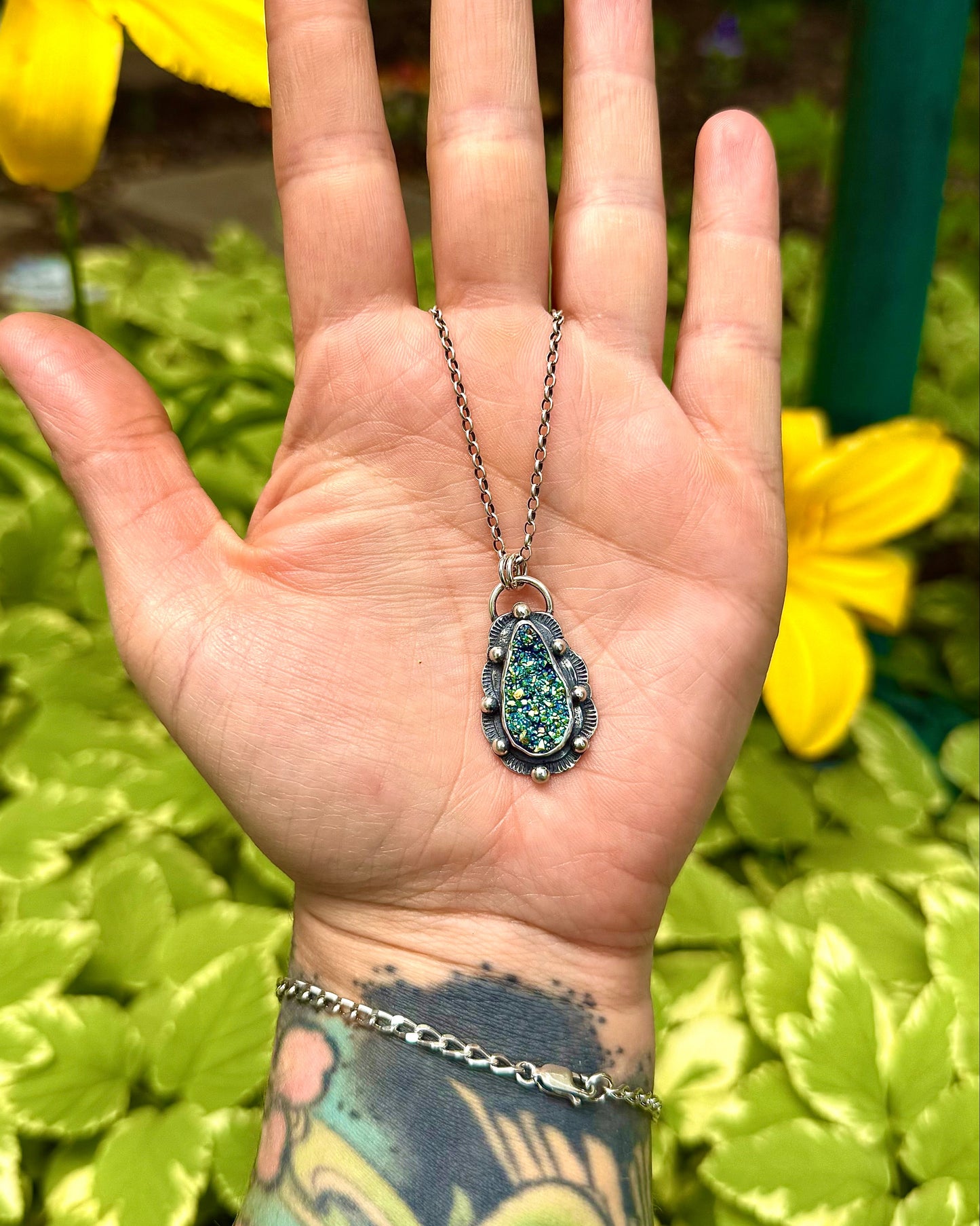 Hand wearing a necklace with a pendant against a green leafy background