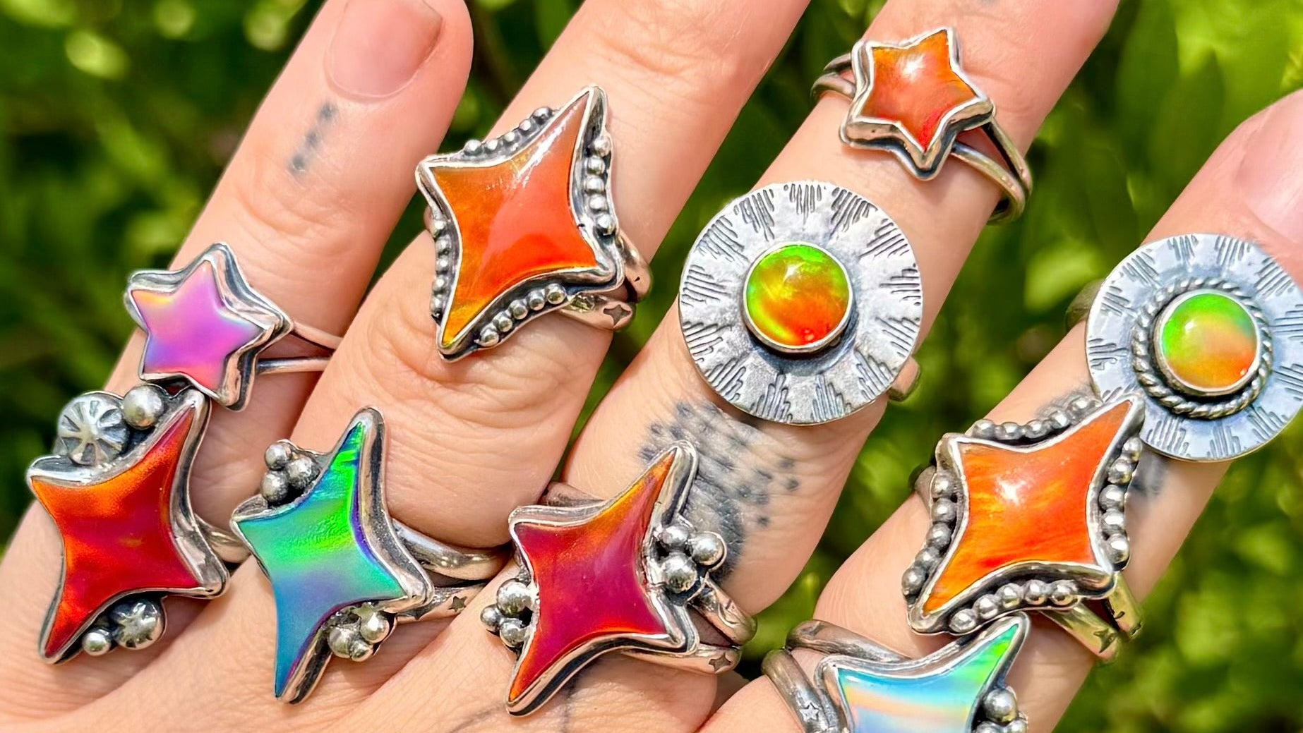 A hand wearing multiple silver rings with colorful star and circular stone designs, photographed outdoors against blurred green foliage.