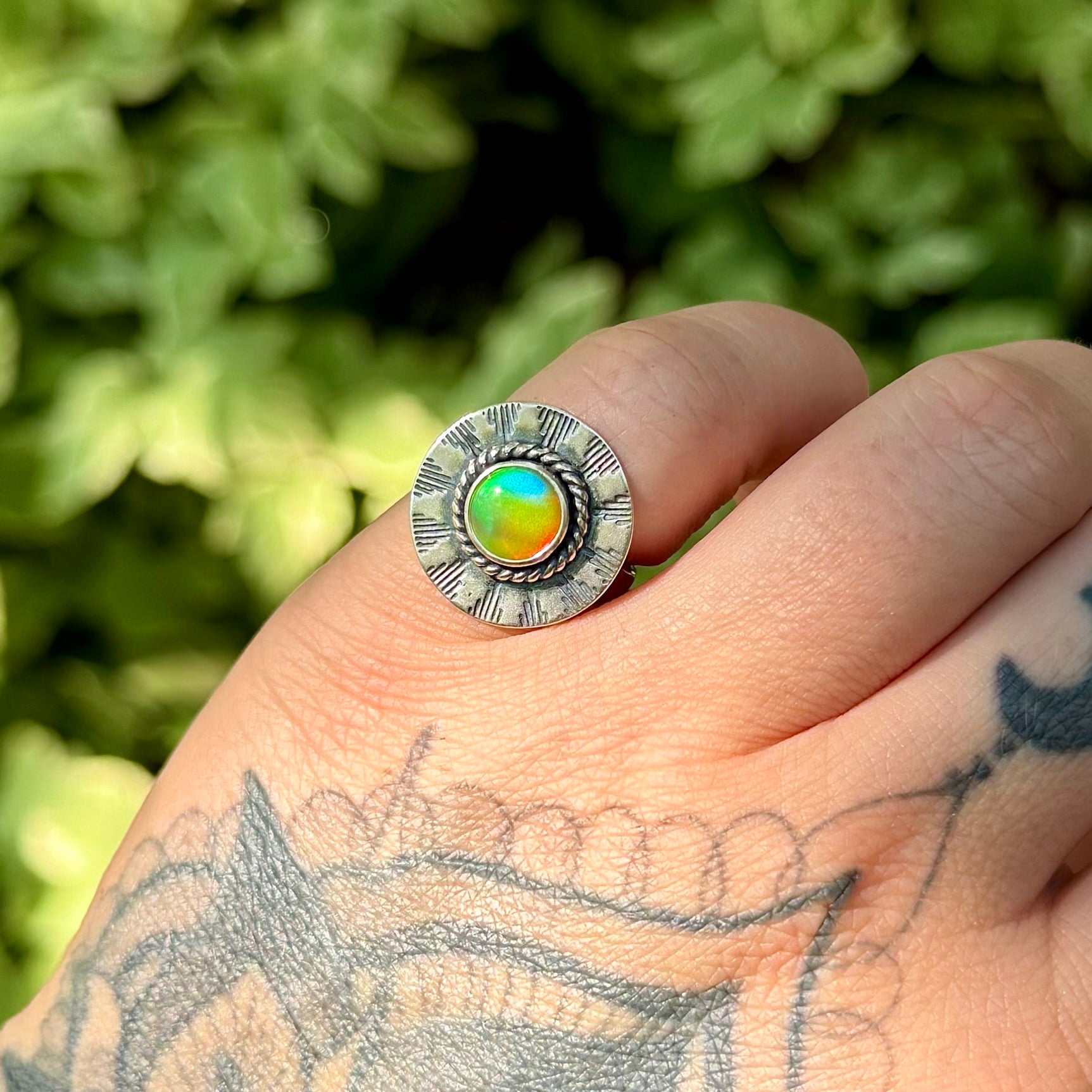 A silver-toned ring with a round, textured bezel holding a vibrant iridescent cabochon gemstone that shifts between green, blue, and orange, worn on a tattooed hand with a blurred green leaf background.