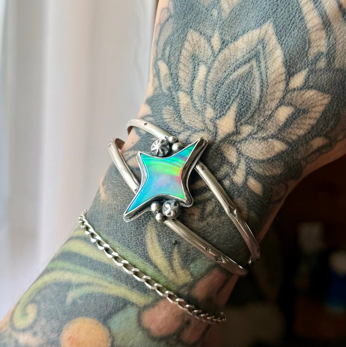 A close-up of a tattooed wrist wearing a silver-toned bangle bracelet with a star-shaped iridescent inlay and small clear stones, along with a chain bracelet.