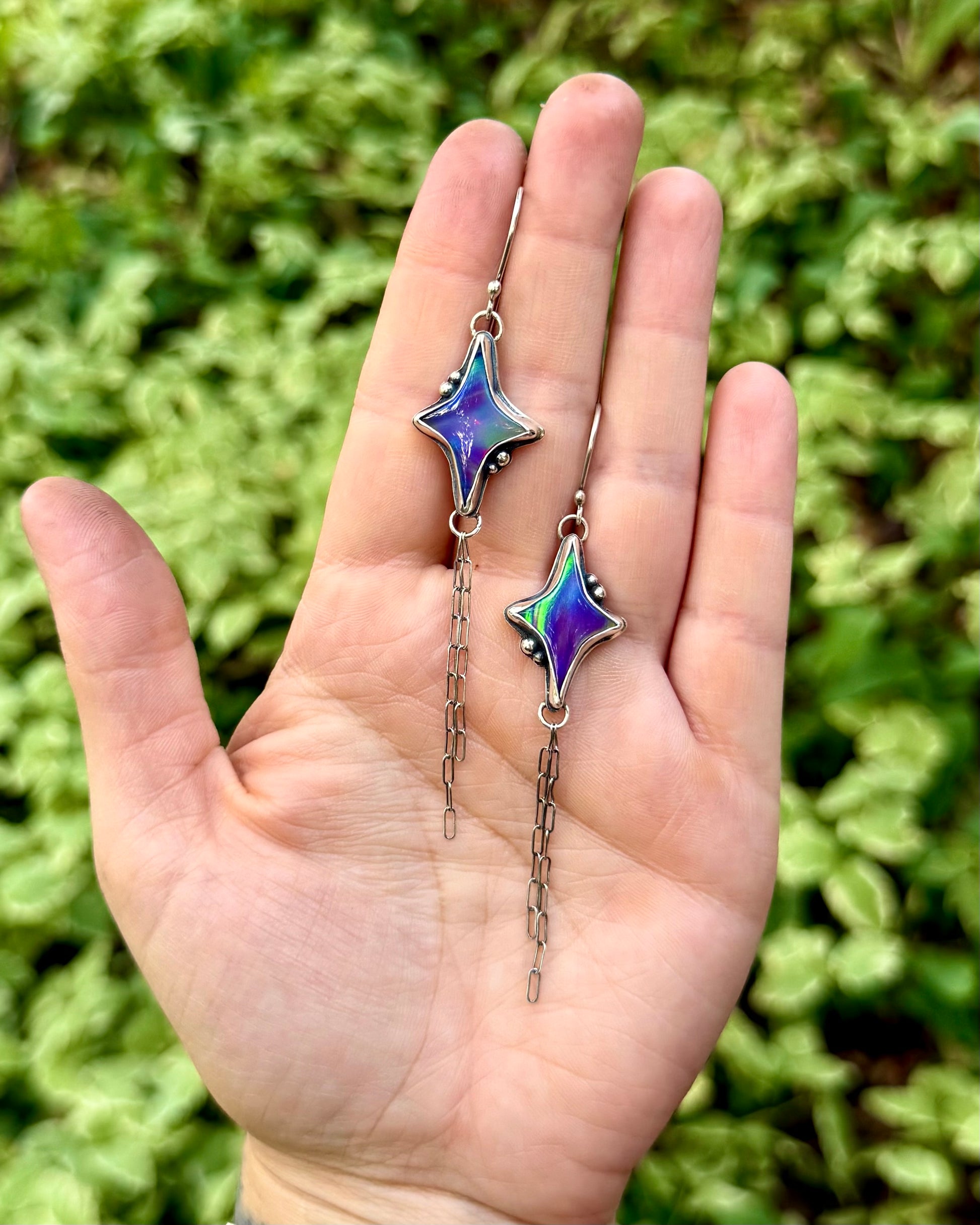 A pair of dangling earrings with star-shaped iridescent purple and blue beads on silver-toned chains, held in a person's hand against a blurred green foliage background.