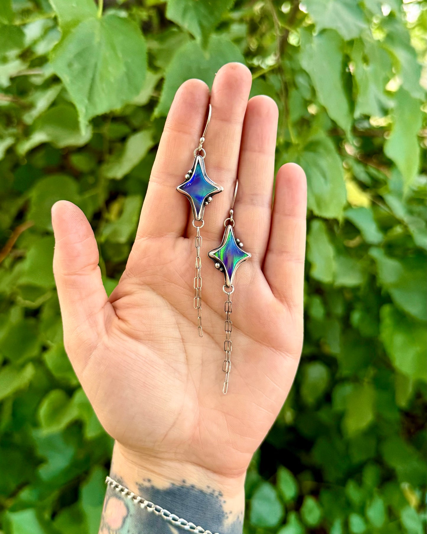 A hand holding a pair of dangling earrings with star-shaped iridescent blue and green charms on silver chains, set against a background of green leaves.