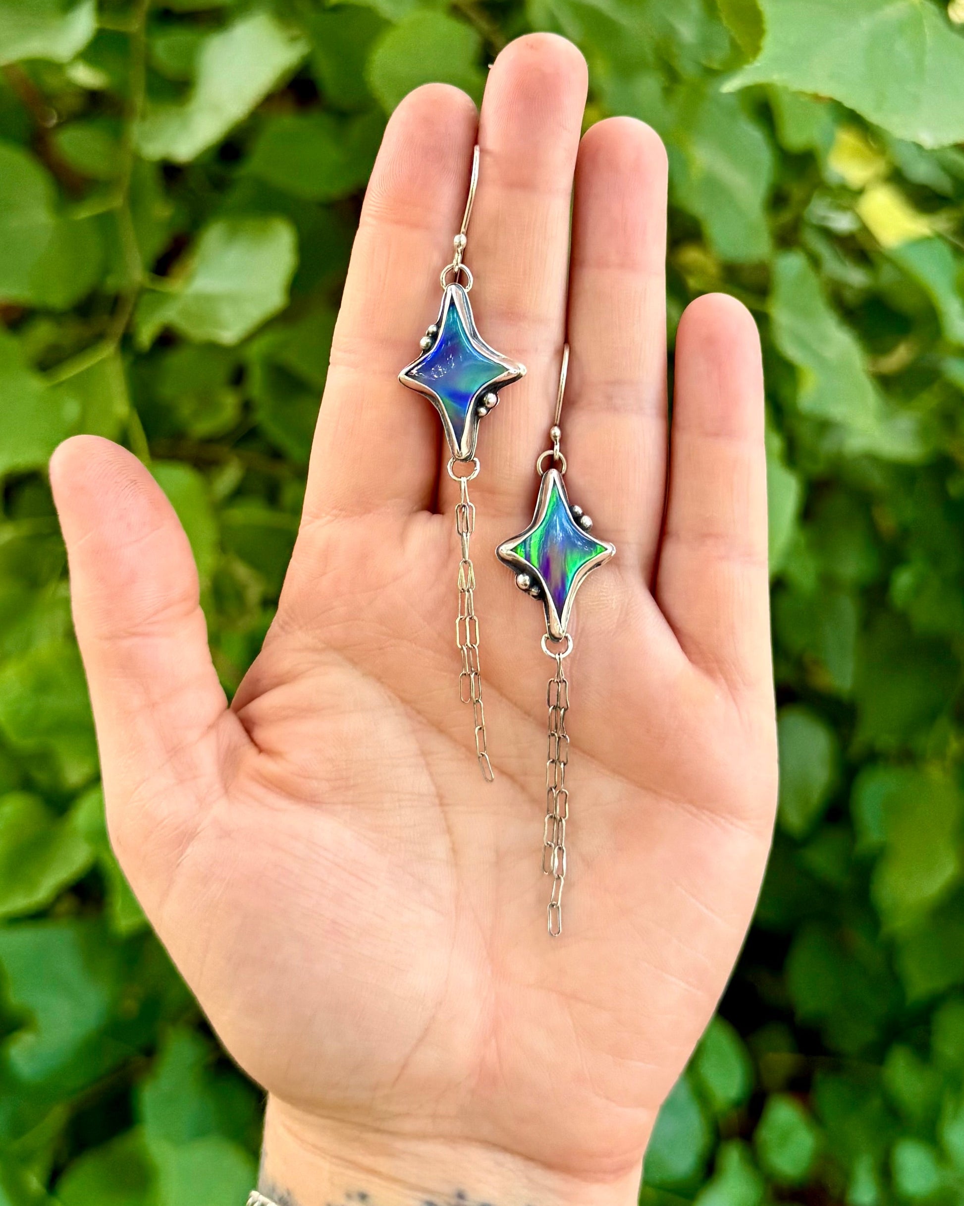 A person's hand holding a pair of dangling earrings with star-shaped iridescent blue and green beads on silver hooks, set against a background of green leaves.