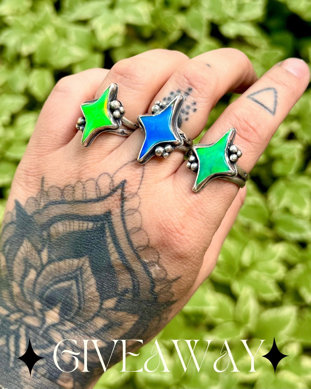 A tattooed hand wearing three star-shaped rings with silver-toned metal and vibrant green, blue, and green enamel inlays, set against a blurred green leaf background with a 'GIVEAWAY' text overlay at the bottom.