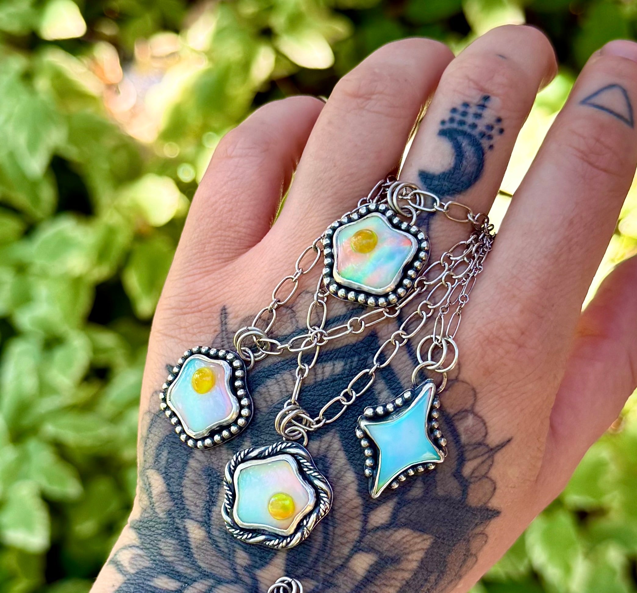 A tattooed hand wearing a silver chain bracelet adorned with multiple iridescent enamel charms, including sunflower, star, and lightning-bolt shapes, set against a blurred green outdoor background.