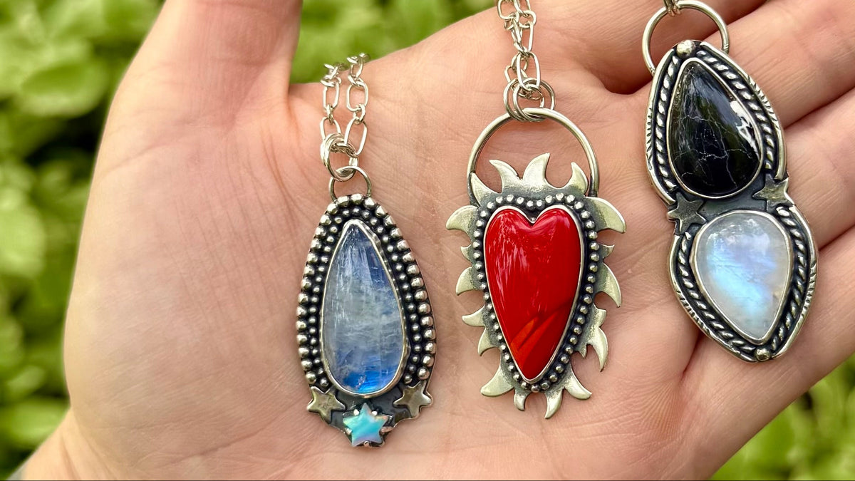 Three silver-toned pendants with teardrop and heart-shaped stones are displayed on an open hand against a blurred green outdoor background. The pendants feature blue, red, black, and milky white stones with decorative silver settings.