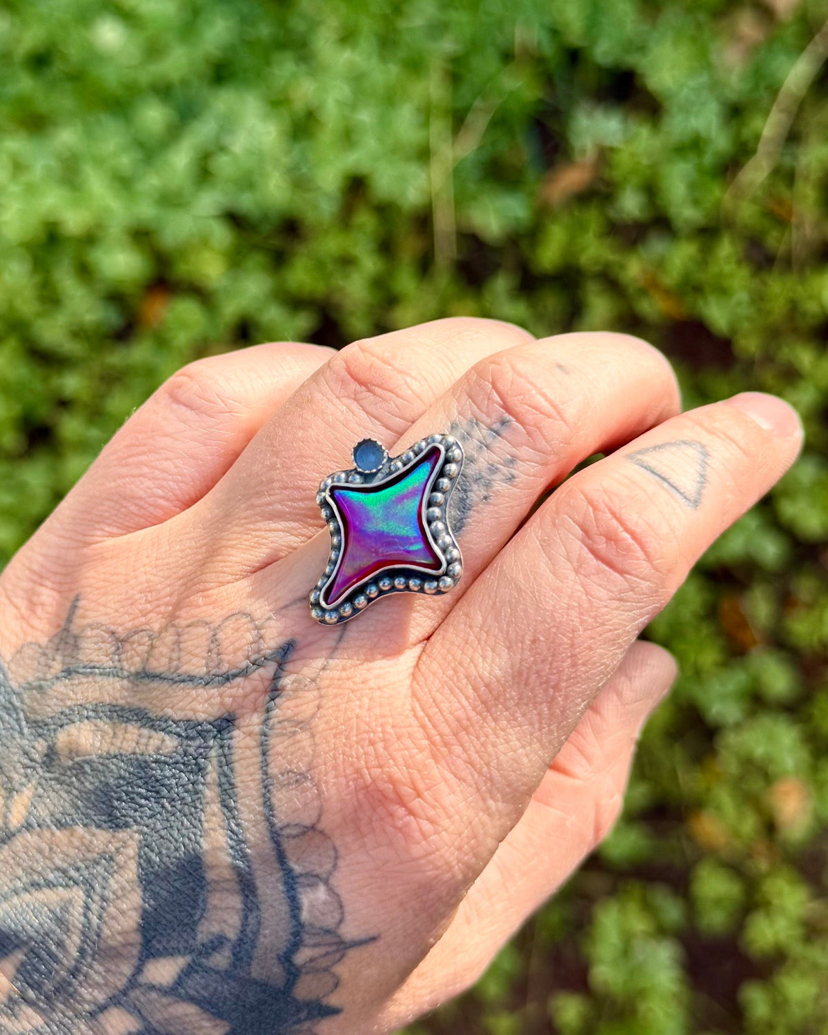 A close-up of a tattooed hand wearing a silver ring with a star-shaped iridescent purple and blue gemstone, set against a blurred green outdoor background.