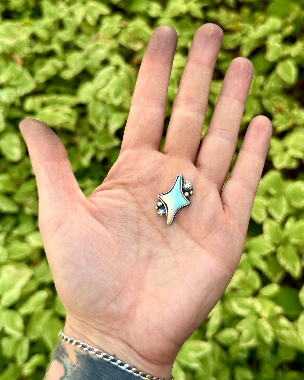 A hand with a silver chain bracelet holds a small silver ring featuring a light blue star-shaped inlay, set against a background of green foliage.