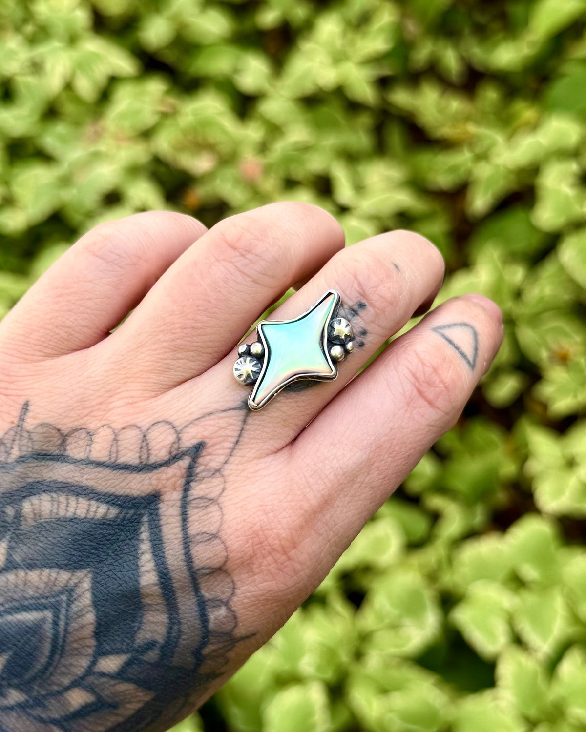 A close-up of a tattooed hand wearing a silver ring with a star-shaped light blue inlay, set against a blurred background of green foliage.