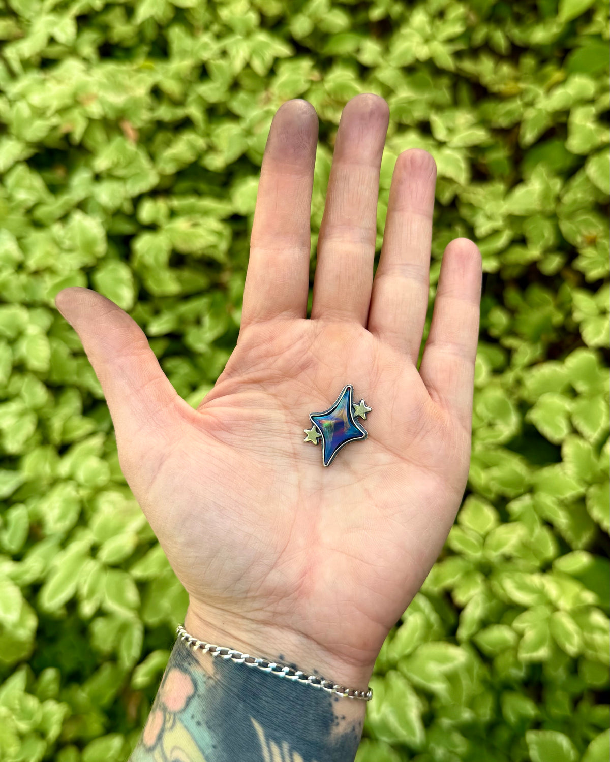 A hand with a tattoo and silver chain bracelet holds a small blue star-shaped pendant with silver details against a background of green leafy plants.