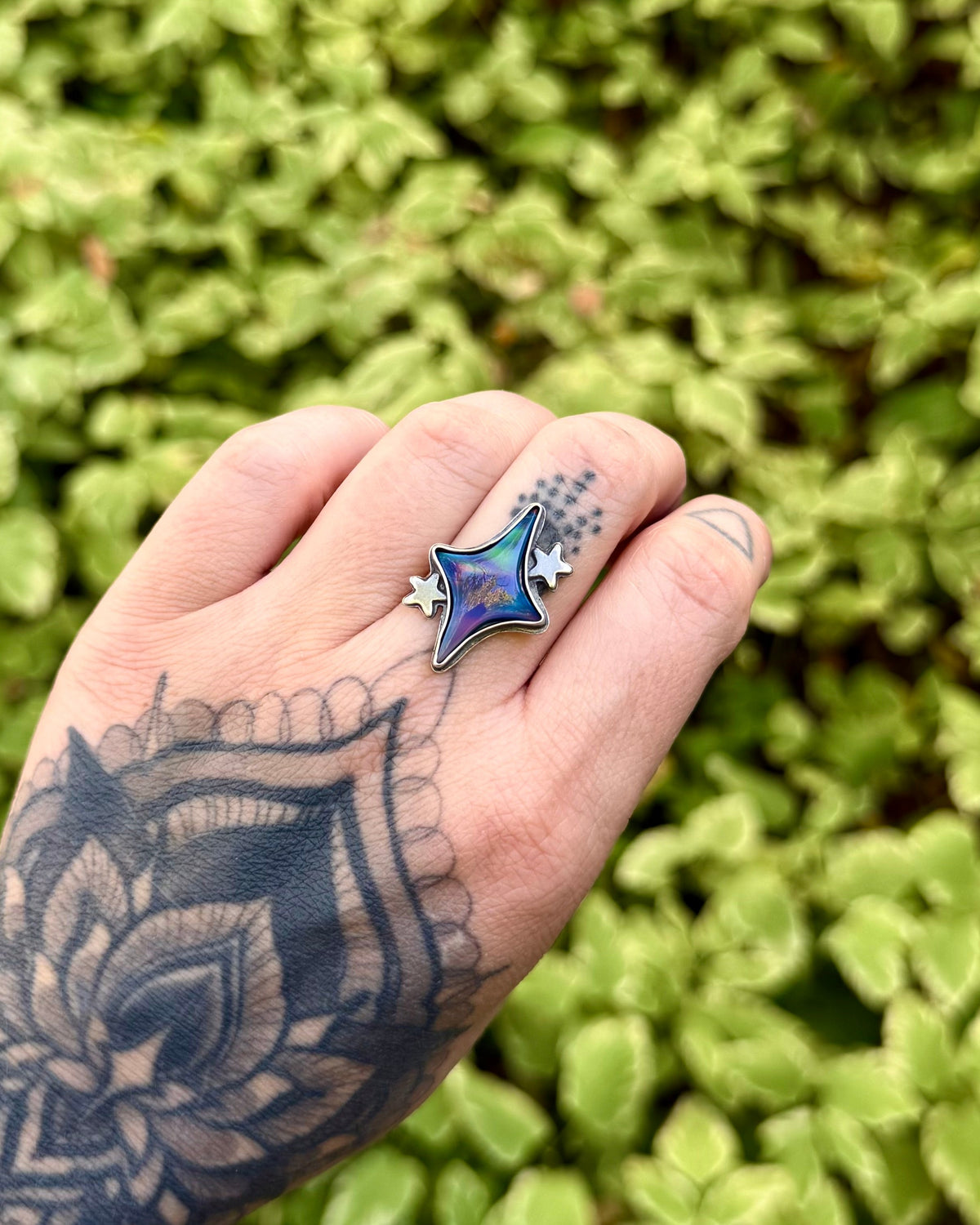 A close-up of a hand with a detailed black mandala tattoo wearing a silver ring with a star-shaped blue and purple inlay, set against a blurred green leaf background.