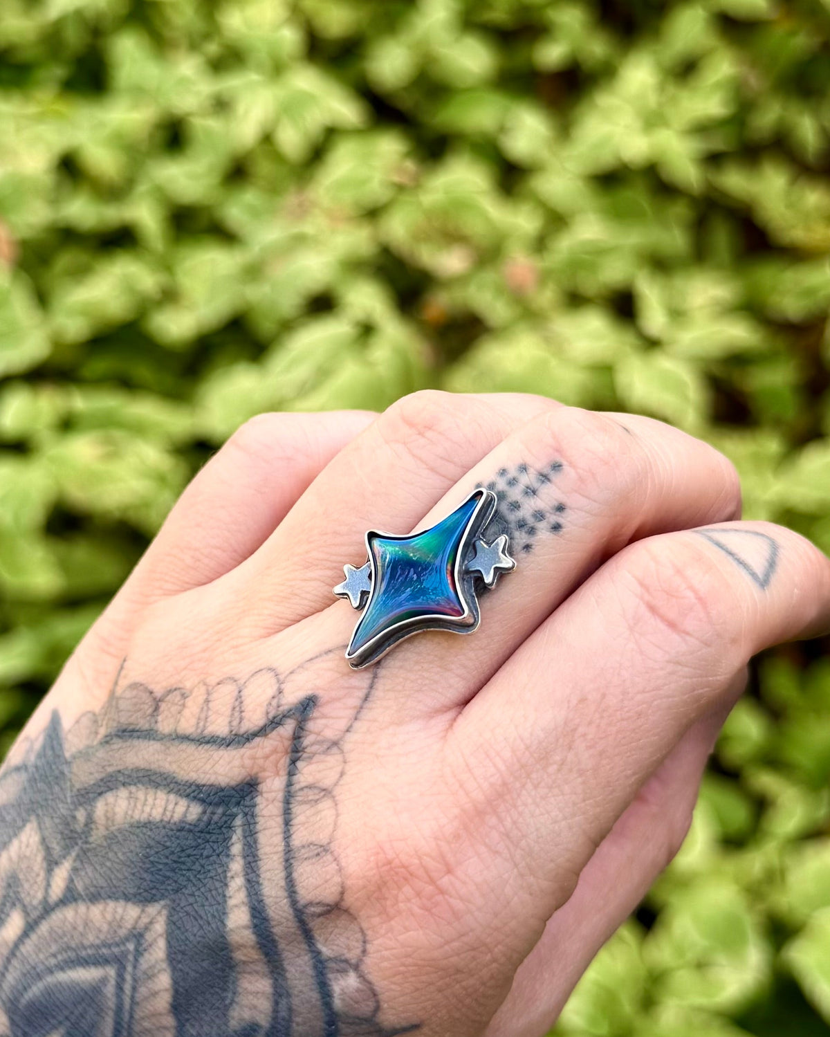 A close-up of a tattooed hand wearing a silver star-shaped ring with a blue inlay, set against a blurred green leaf background.