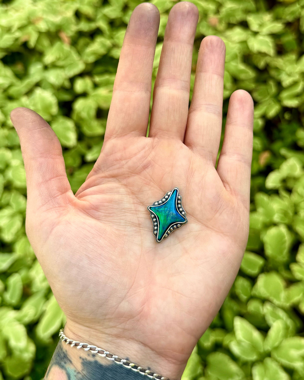 A small star-shaped turquoise and blue bead pendant with silver trim is held in the center of an open hand, with a blurred green leafy background behind it.