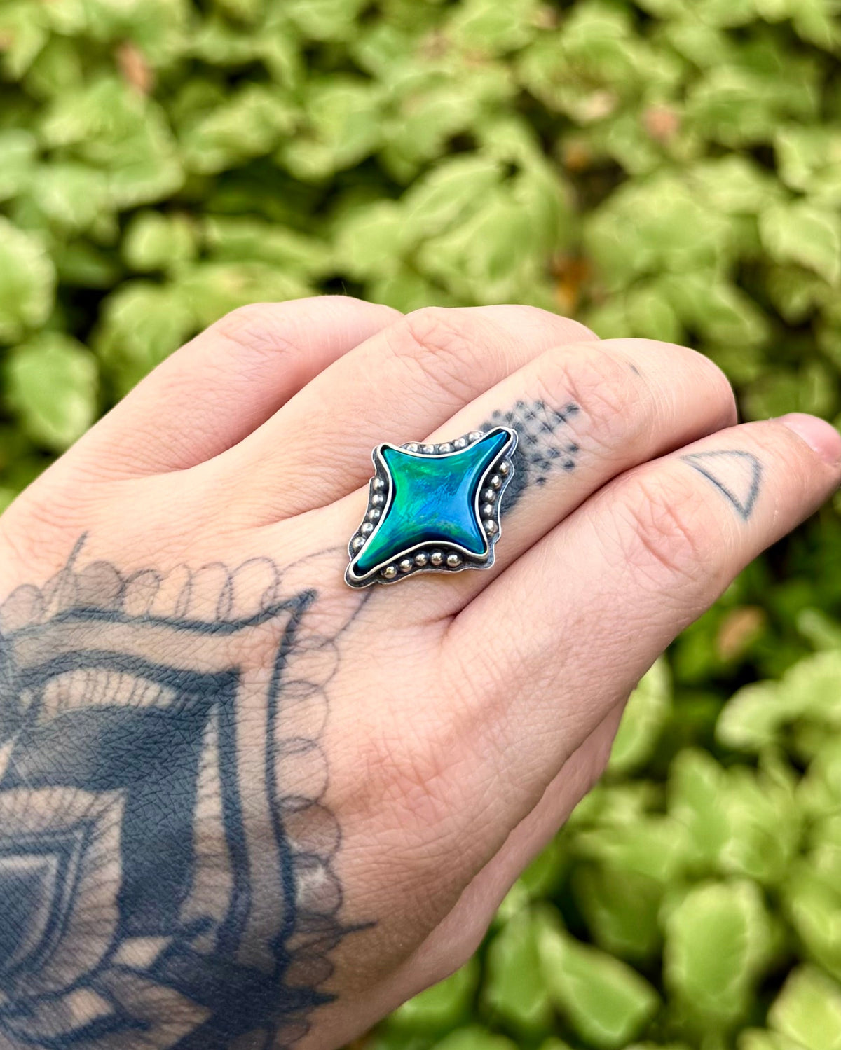 A hand with intricate black tattoos wears a turquoise and black geometric star-shaped ring, set against a blurred background of green foliage.