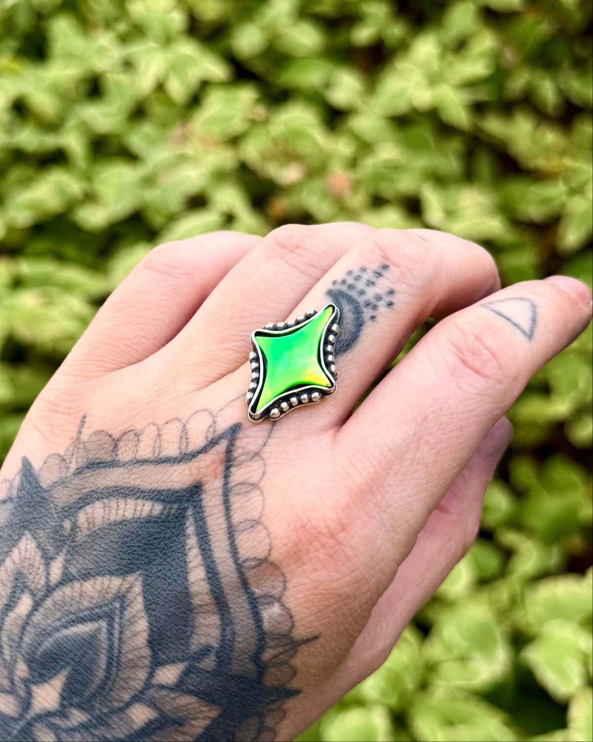 A close-up of a hand with black tattoos wearing a silver ring with a bright green star-shaped stone, set against a blurred green leaf background.