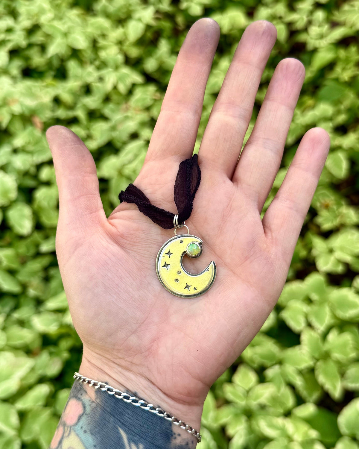 A hand holding a yellow crescent moon pendant necklace with a small green gemstone and black ribbon, set against a background of green foliage.