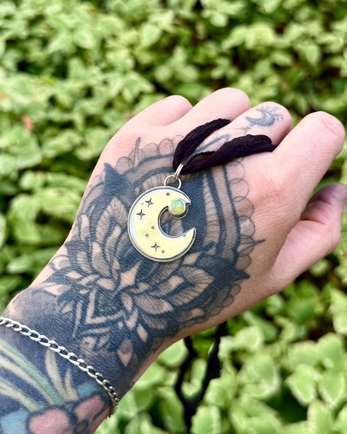 A close-up of a hand with intricate black floral tattoos holding a crescent moon pendant necklace. The pendant has a pale yellow-green enamel face with small gold stars and a central green gemstone, suspended from a dark brown cord.