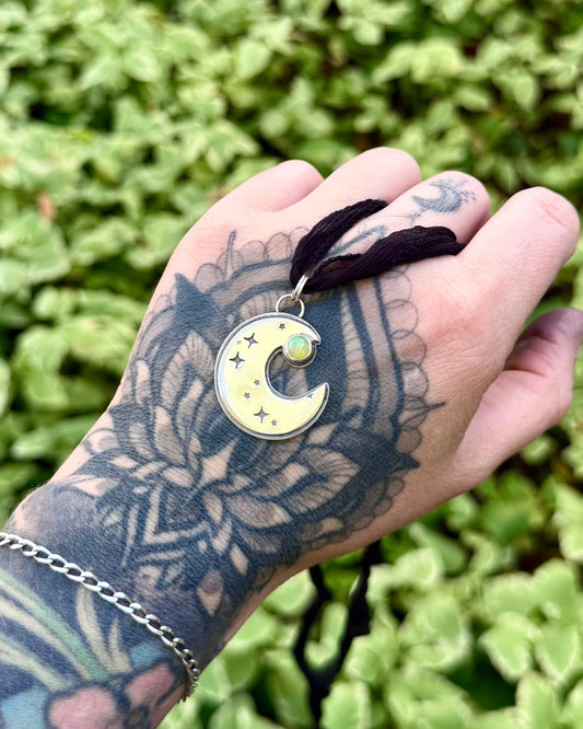 A close-up of a hand with intricate black floral tattoos holding a crescent moon pendant necklace. The pendant has a pale yellow-green enamel face with small gold stars and a central green gemstone, suspended from a dark brown cord.