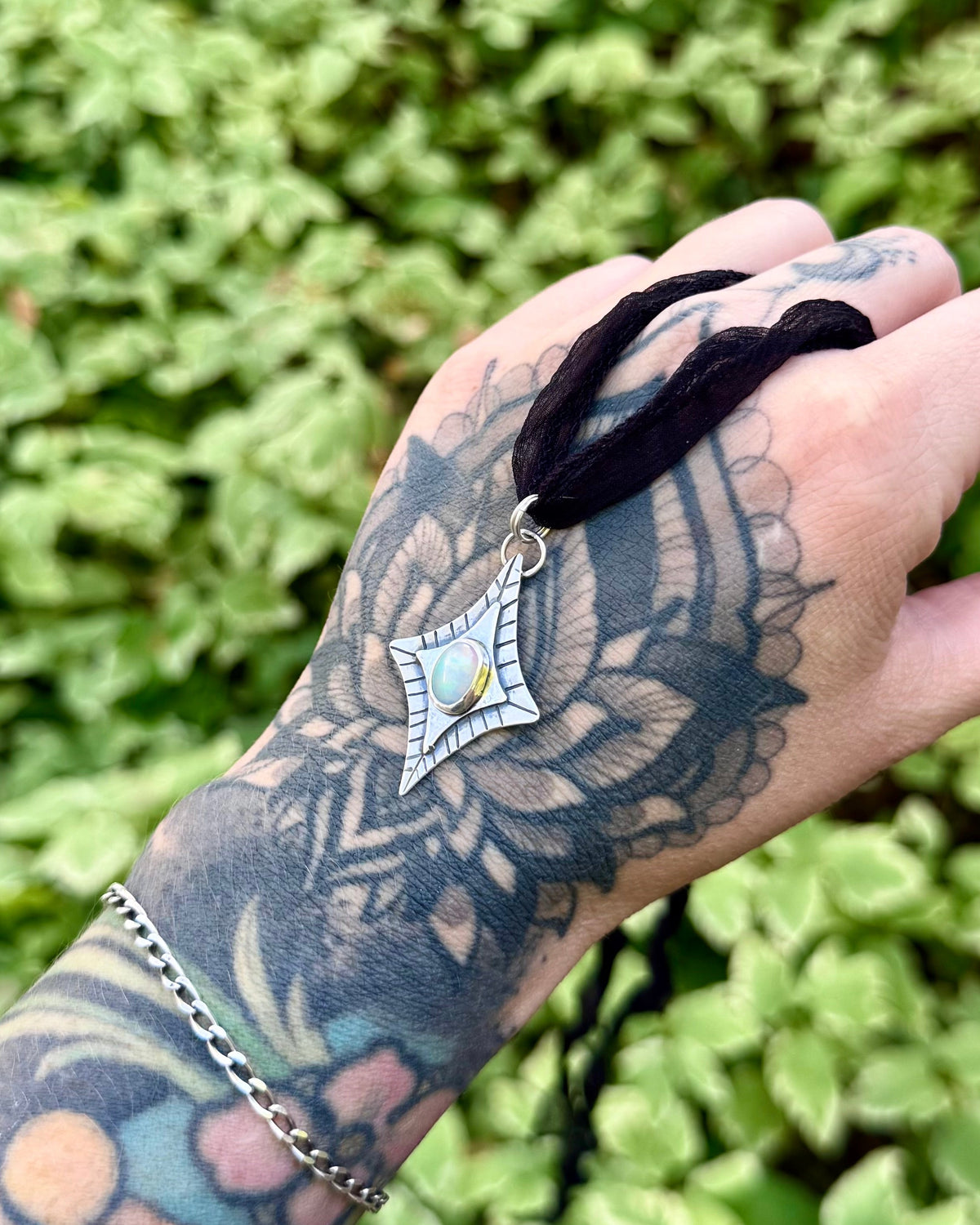A close-up of a tattooed hand holding a black cord necklace with a silver-toned pendant featuring a blue and white star-like design, set against a blurred green outdoor background.