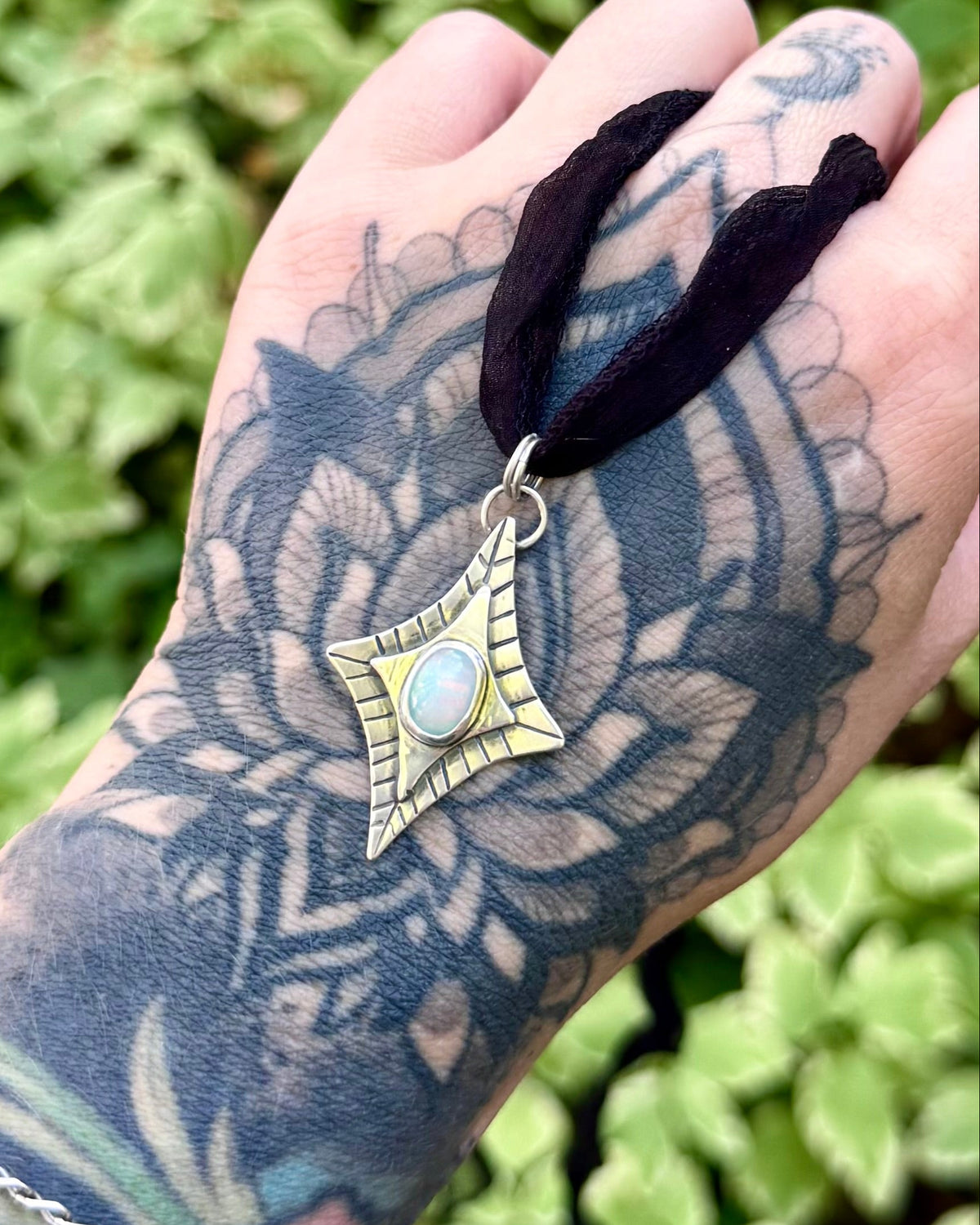 A tattooed hand holding a black cord necklace with a gold-toned pendant featuring a book and a small bird, set against a blurred background of green foliage.