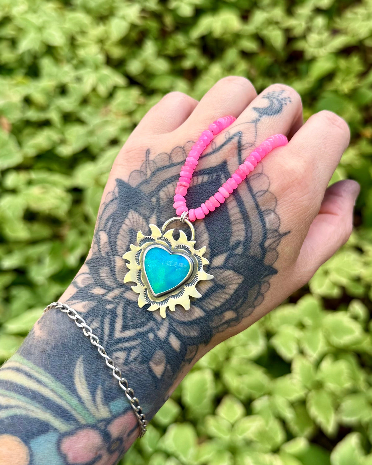 A tattooed hand holding a pink beaded necklace with a turquoise heart pendant against a background of green foliage.